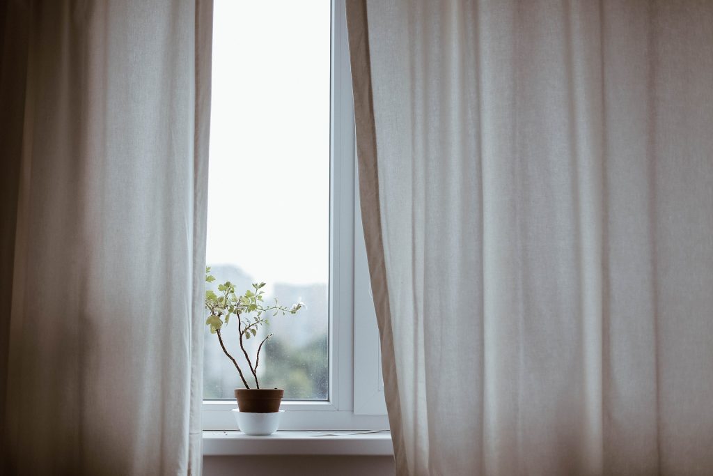 Une fenêtre sur laquelle des voilages beiges sont posés et entre lesquels on peut voir un pot de fleurs