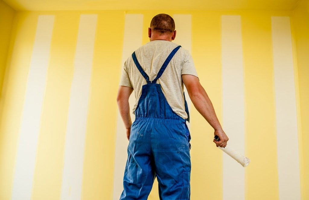 homme de dos en bleu de travail qui applique de la peinture jaune